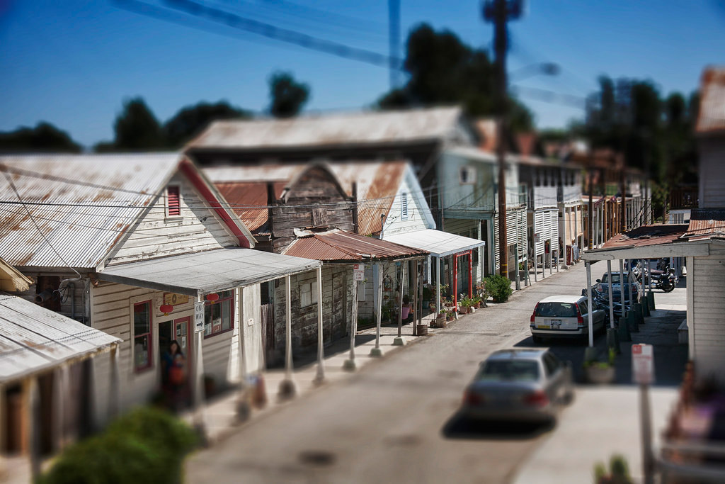 A row of beaten-down houses on a one way street, very close together. Cars are parked on one side of the street. The edges are blurred, the sky is bright blue.