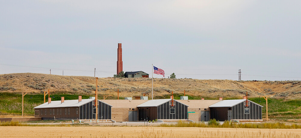 A view of the Heart Mountain Interpretive Center, built to look like three World War II Japanese American incarceration barracks. On a hill in the background, the old hospital building with its iconic red chimney overlooks the Interpretive Center.