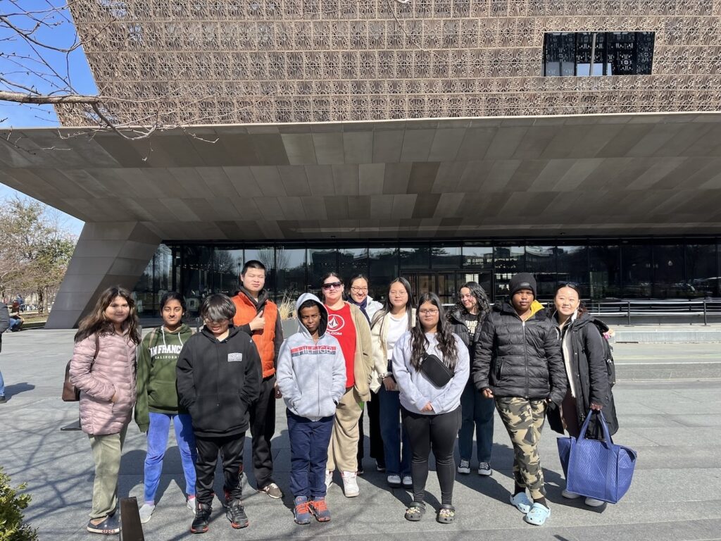 A group of youth wearing winter attire stand in front of the National Museum of African American History and Culture.